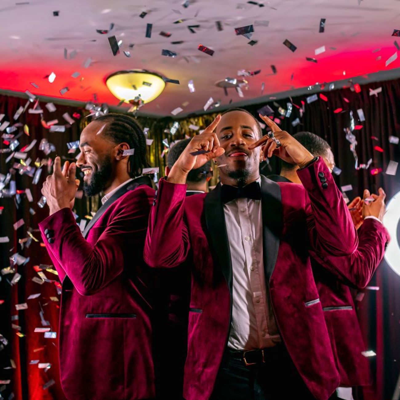 Two guests in burgundy tuxedos posing as silver confetti falls around the 360 video booth