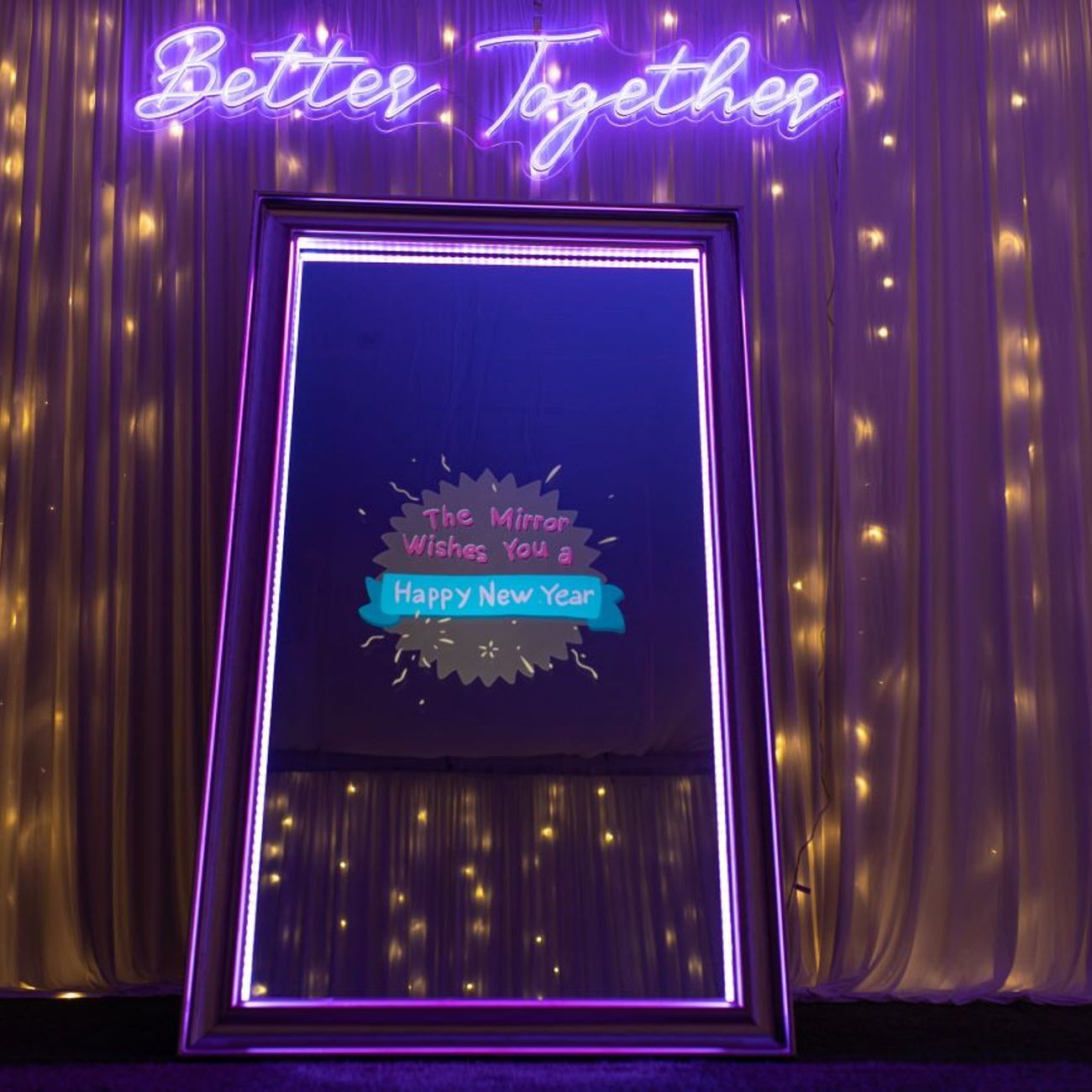 Full-length magic mirror photo booth in front of a fairy-light curtain backdrop, neon 'Better Together' sign overhead, mirror screen showing a 'Happy New Year' animated splash