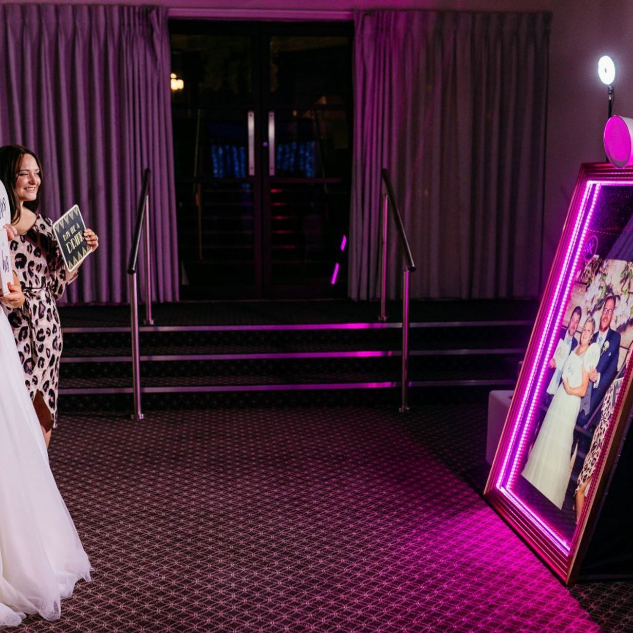Bride, groom and bridesmaids posing in front of the magic mirror at a hotel wedding, hot-pink LED frame around the mirror, ring light on top and the group preview showing on the mirror's touchscreen