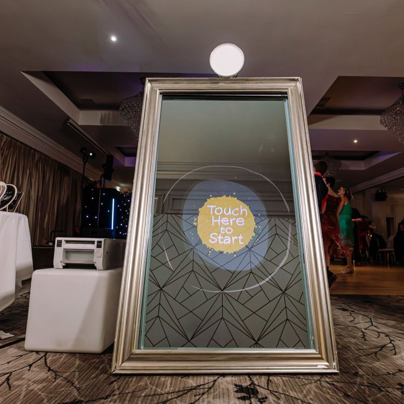 Silver-framed full-length magic mirror set up in a country-house hotel ballroom, 'Touch Here to Start' prompt on the screen, dye-sublimation printer on a side table and stacked photo albums beside it