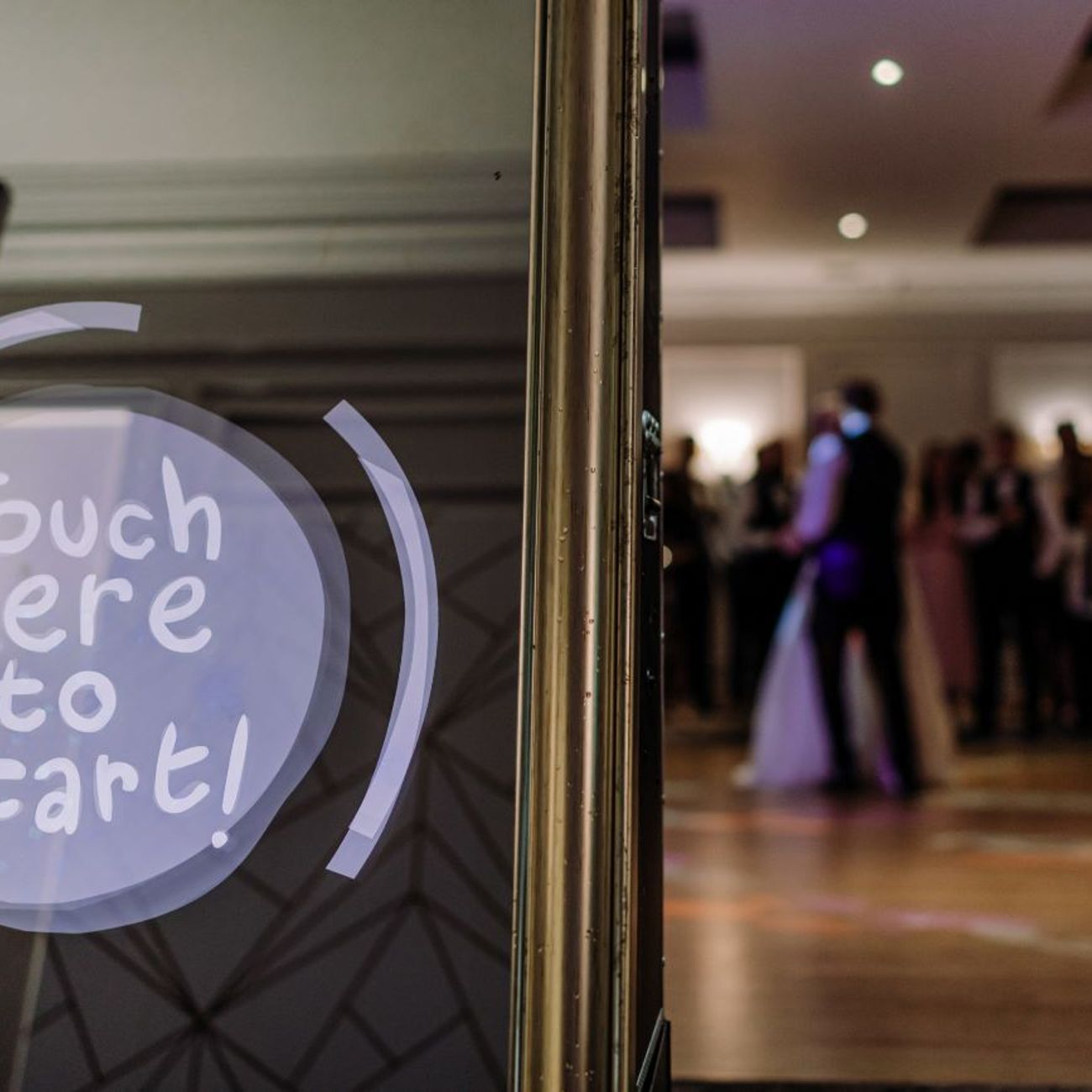 Close-up of the magic mirror's 'Touch Here to Start' animated prompt, with the bride, groom and wedding guests gathered on the dance floor reflected in the mirror behind
