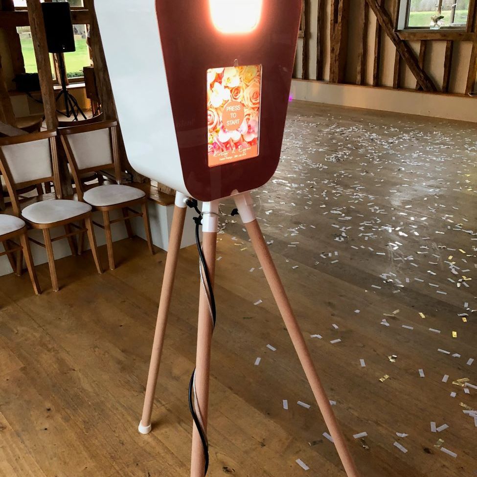 Retro pod set up in a rustic barn wedding venue with confetti scattered across the wooden floor, touchscreen showing a rose-floral 'Press to Start' overlay branded for the couple