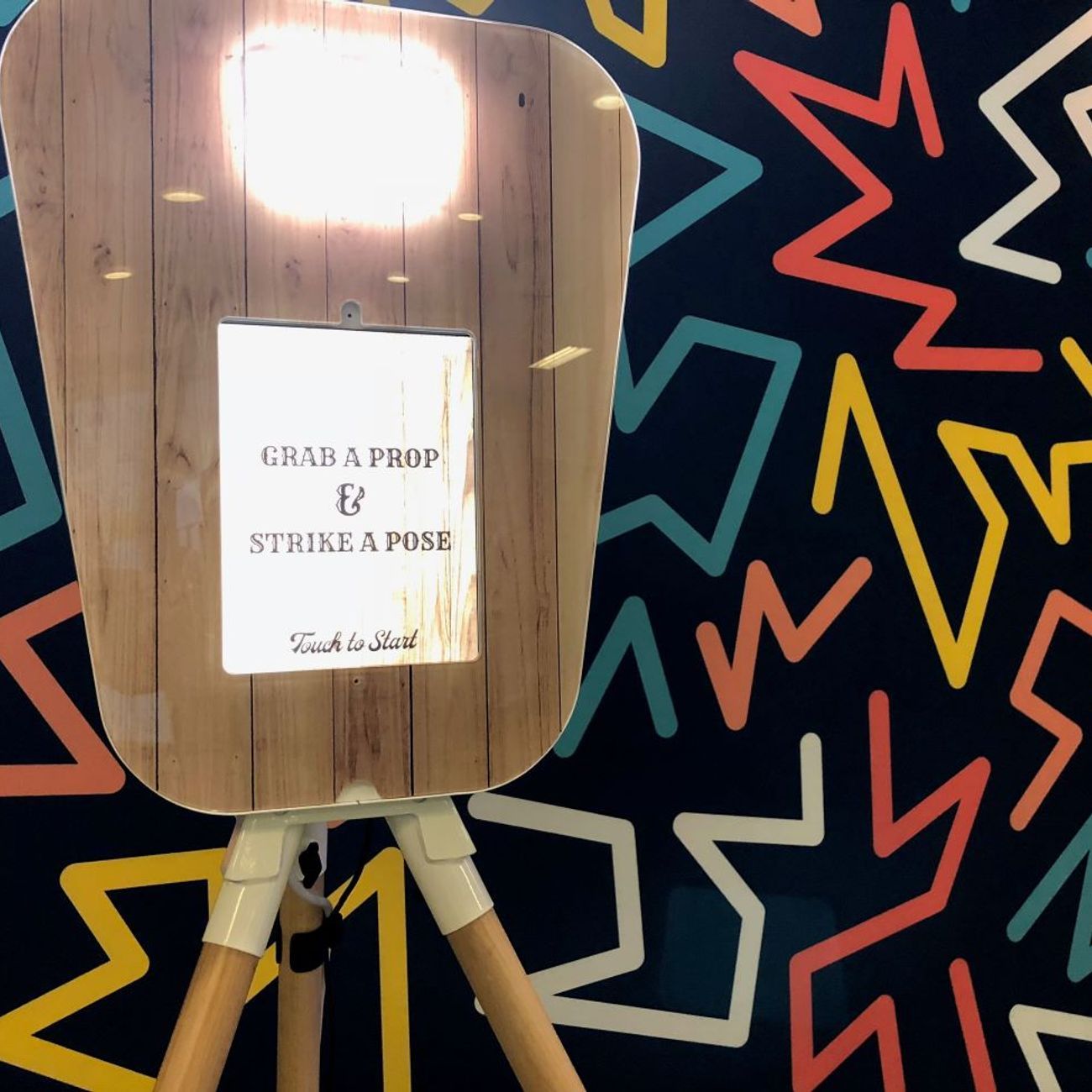 Front of the retro pod's wood-fascia panel — fill light at the top, 'Grab a Prop & Strike a Pose / Touch to Start' touchscreen graphic, against the Memphis-style geometric backdrop