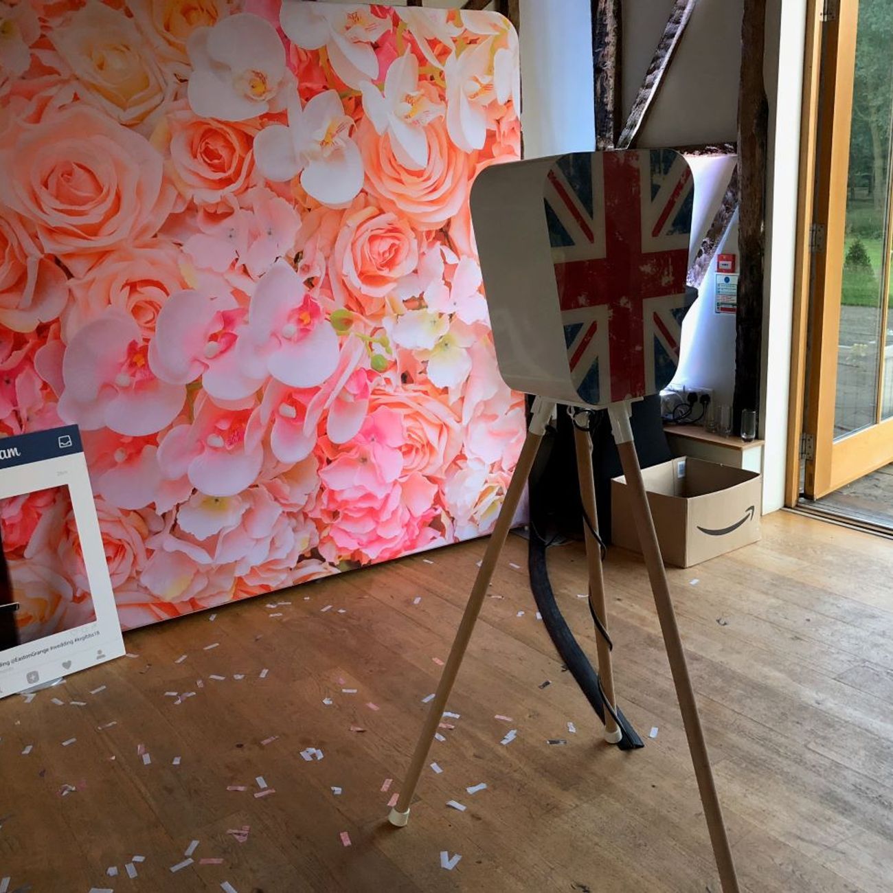 Retro pod on its wooden tripod next to a pink-and-white flower wall backdrop in a barn venue, with an Instagram-frame prop and confetti scattered on the floor