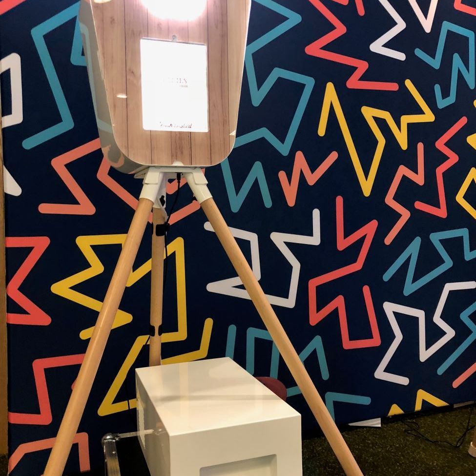 Retro pod photo booth on its wooden tripod against a Memphis-style geometric backdrop in navy with red, yellow, blue and white chevrons, fill light illuminated, separate white printer flight case on the floor in front