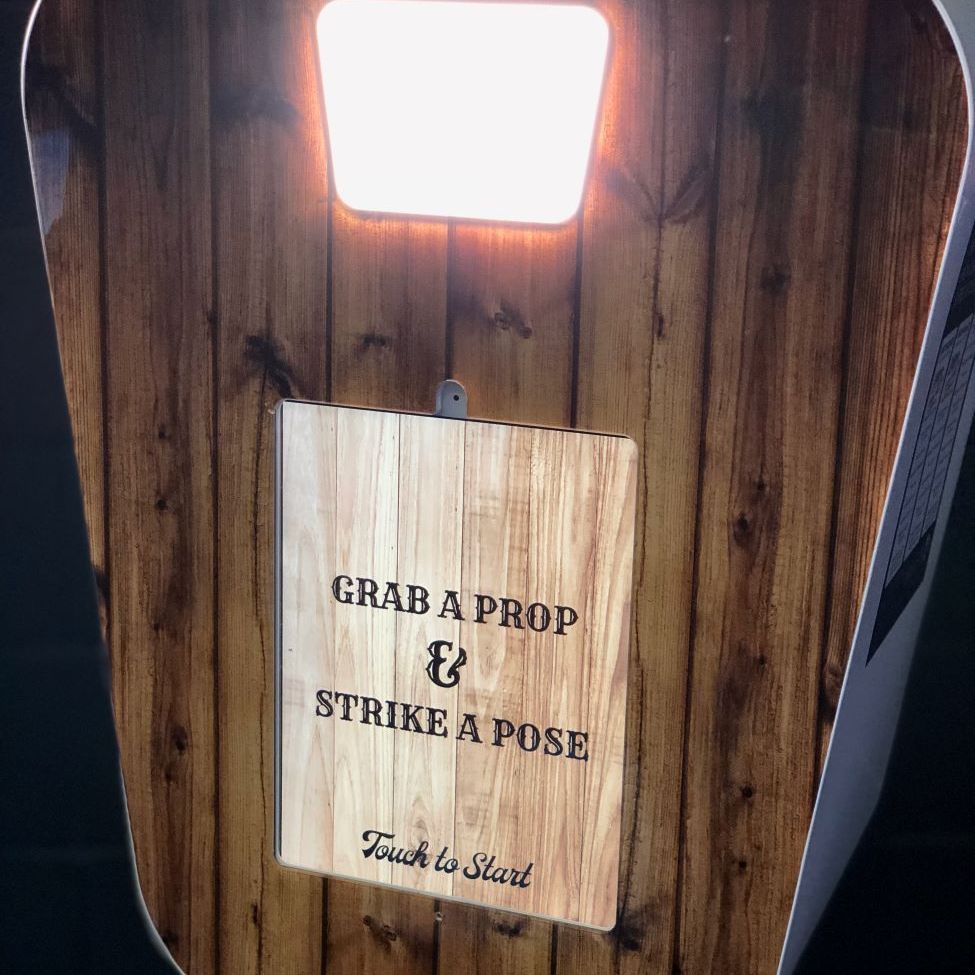 Tight detail of the retro pod's wood-grain fascia — fill-light panel at the top, central wood-pattern screen reading 'Grab a Prop & Strike a Pose / Touch to Start'