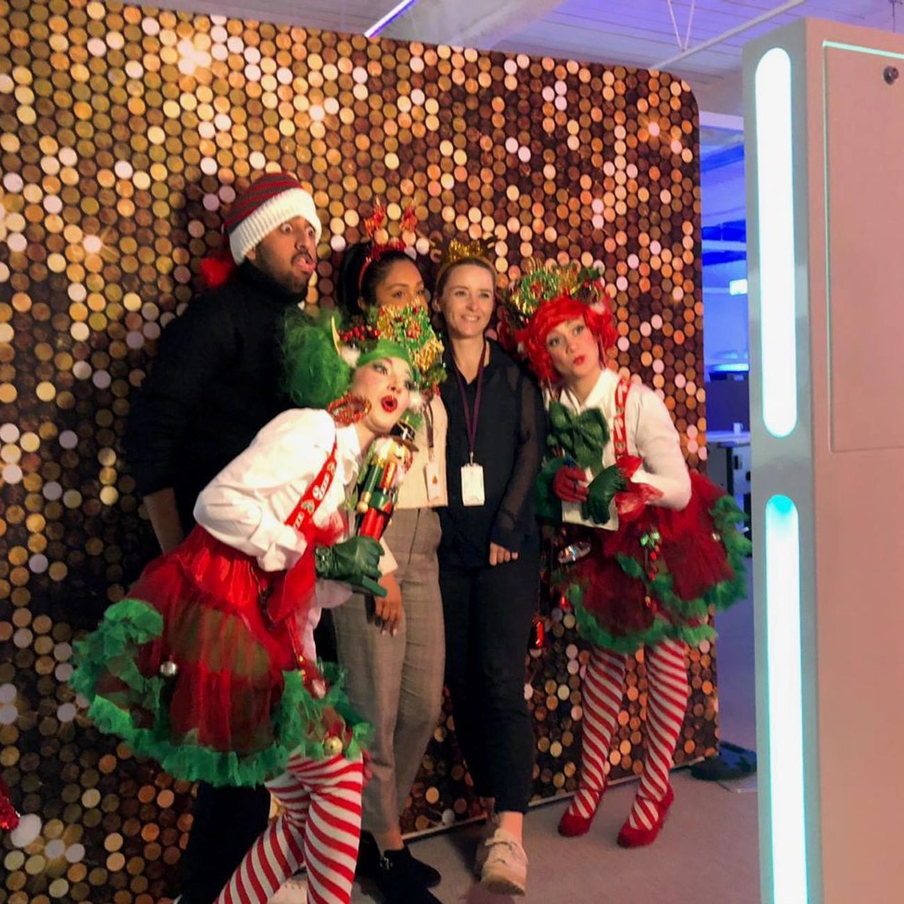 Corporate Christmas group photo at the selfie pod — guests in elf and nutcracker costumes posing in front of a gold sequin backdrop