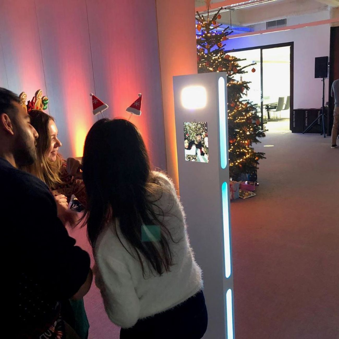 Three guests in Christmas reindeer and Santa hat headbands using the selfie pod at an office Christmas party, group preview showing on the touchscreen
