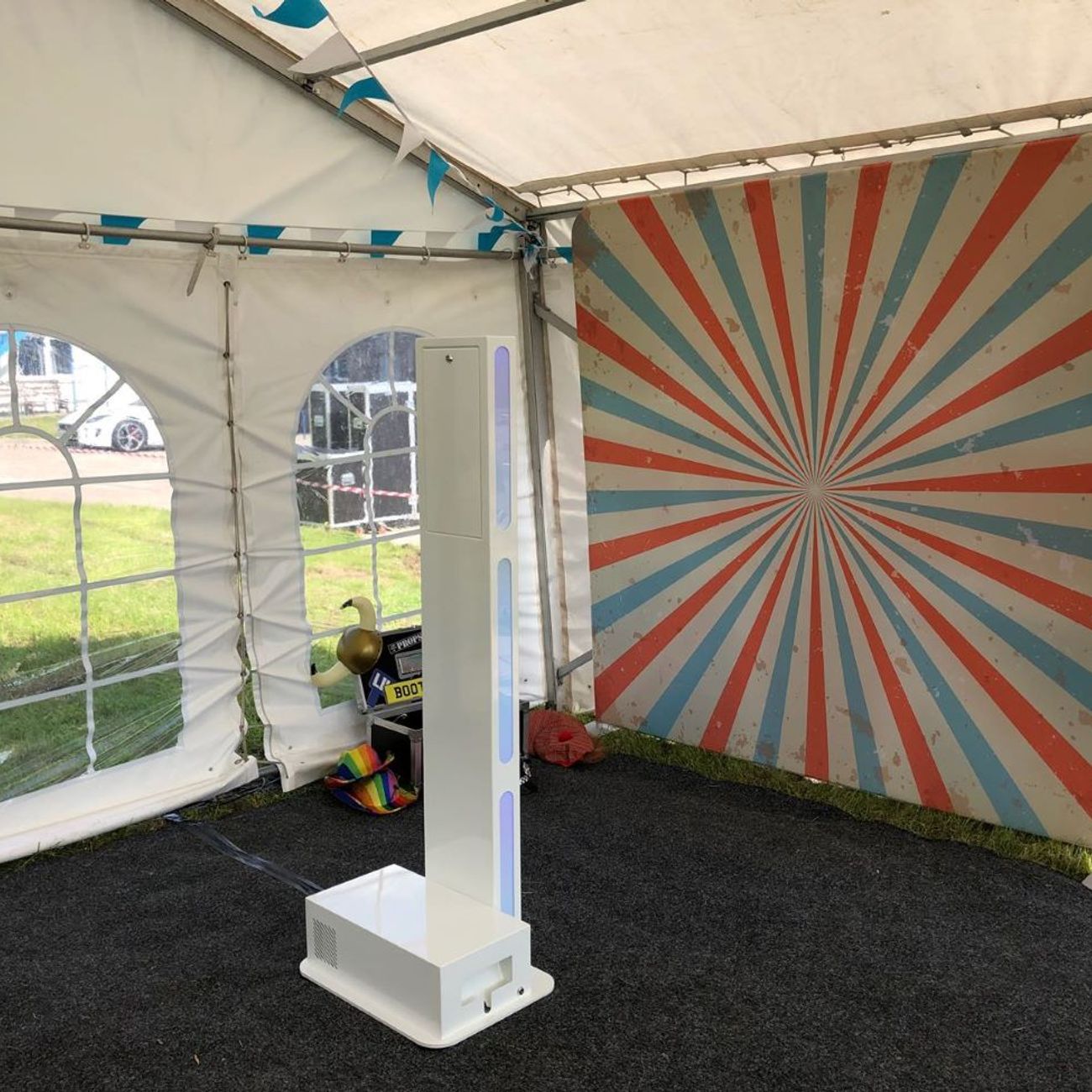 Selfie pod set up inside a garden marquee next to a red-and-cream sunburst backdrop, slim footprint visible against the marquee window