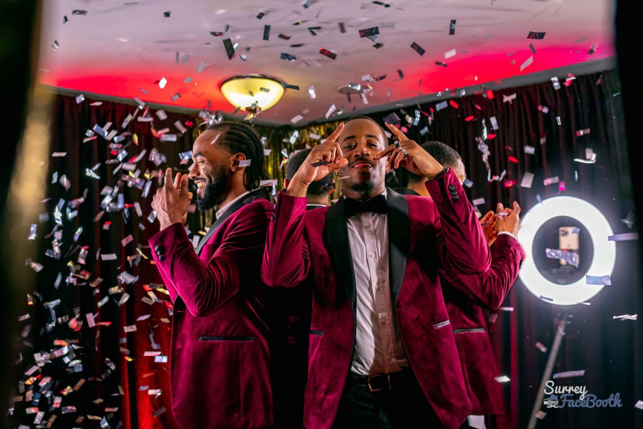 Two guests in burgundy tuxedos posing as silver confetti falls around the 360 video booth