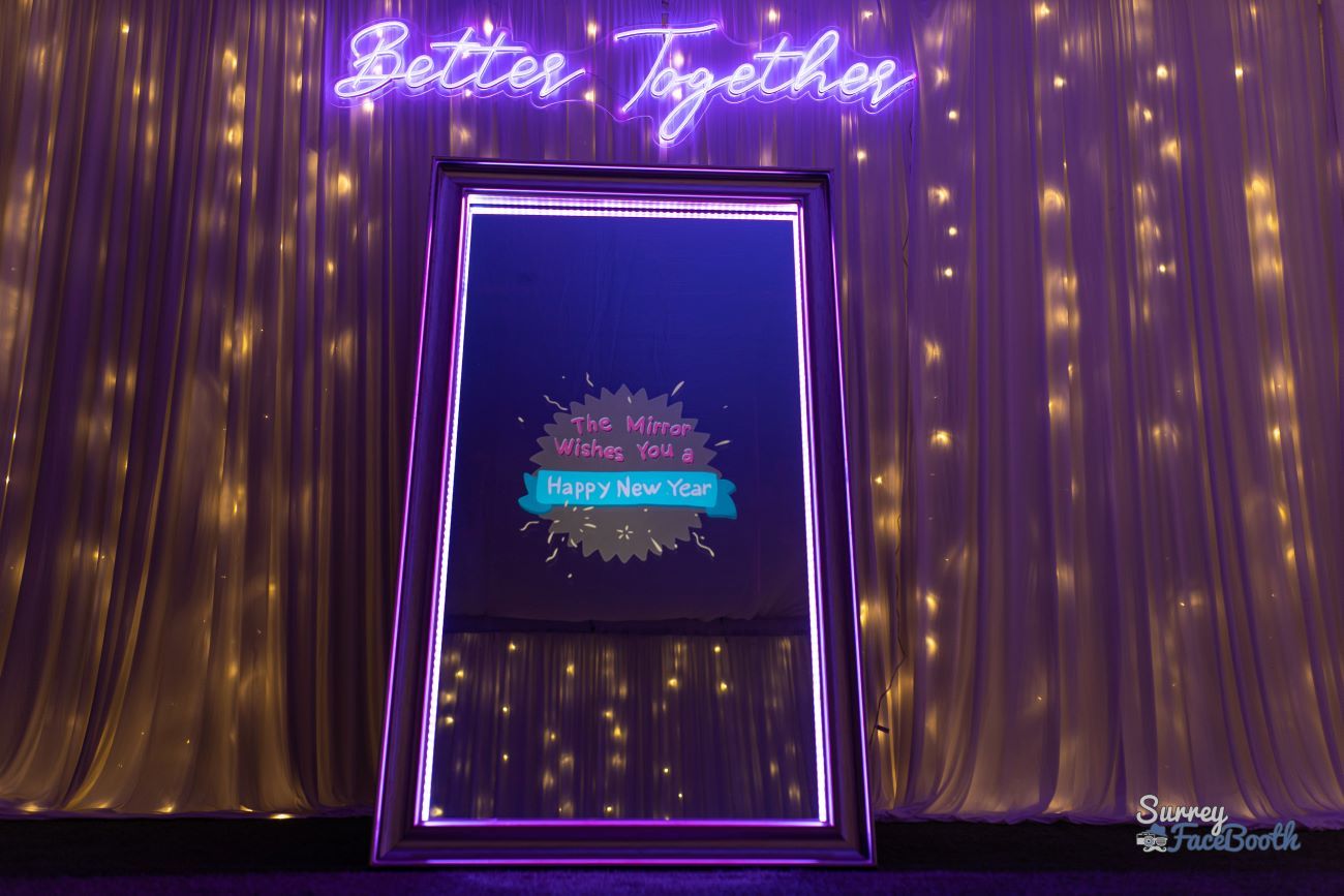 Full-length magic mirror photo booth in front of a fairy-light curtain backdrop, neon 'Better Together' sign overhead, mirror screen showing a 'Happy New Year' animated splash