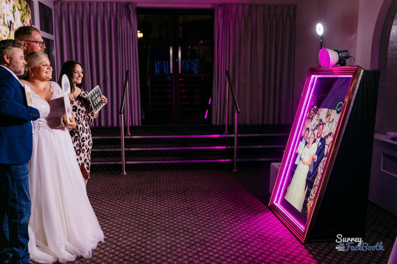 Bride, groom and bridesmaids posing in front of the magic mirror at a hotel wedding, hot-pink LED frame around the mirror, ring light on top and the group preview showing on the mirror's touchscreen