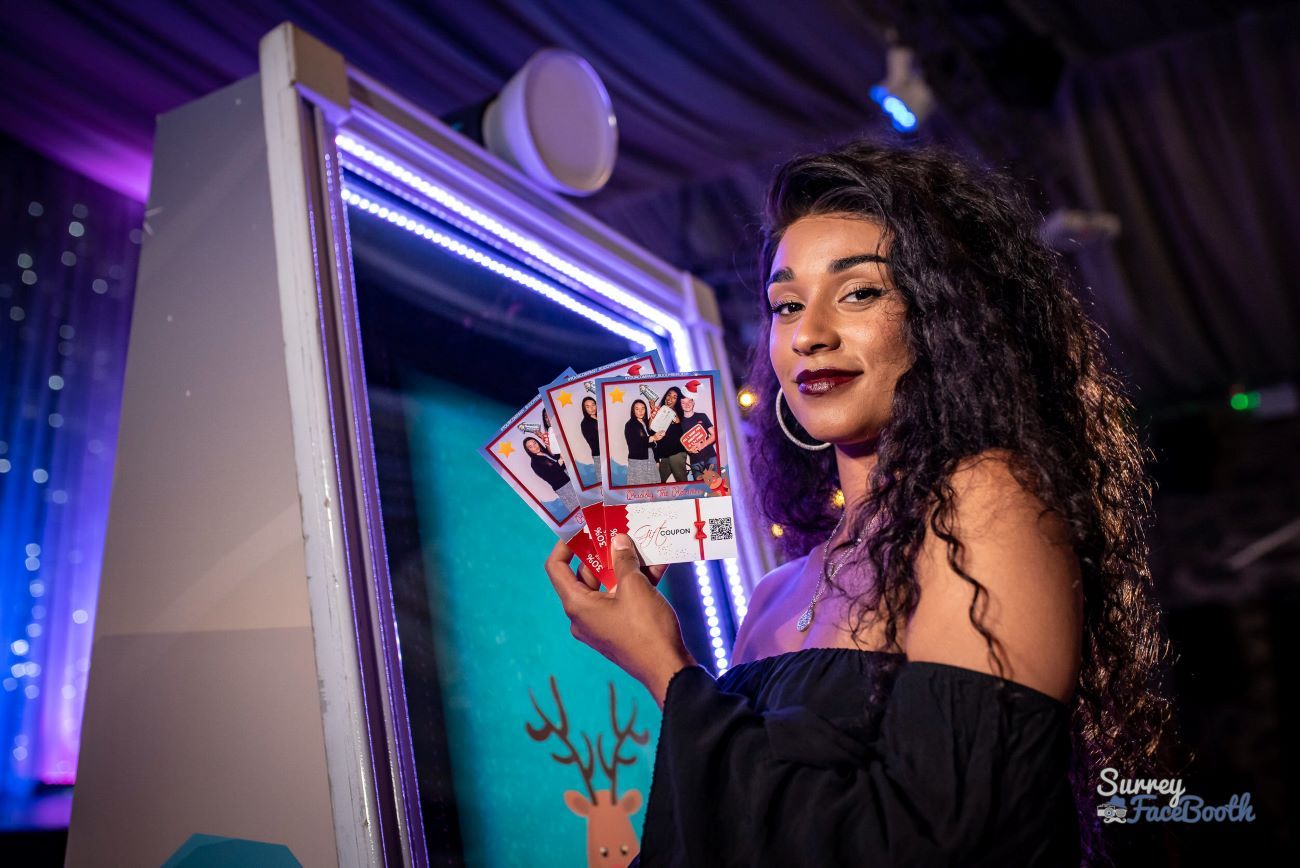 Guest holding up four custom-branded photo prints next to the magic mirror at a Christmas event, LED frame illuminated and dye-sublimation printer visible on the booth