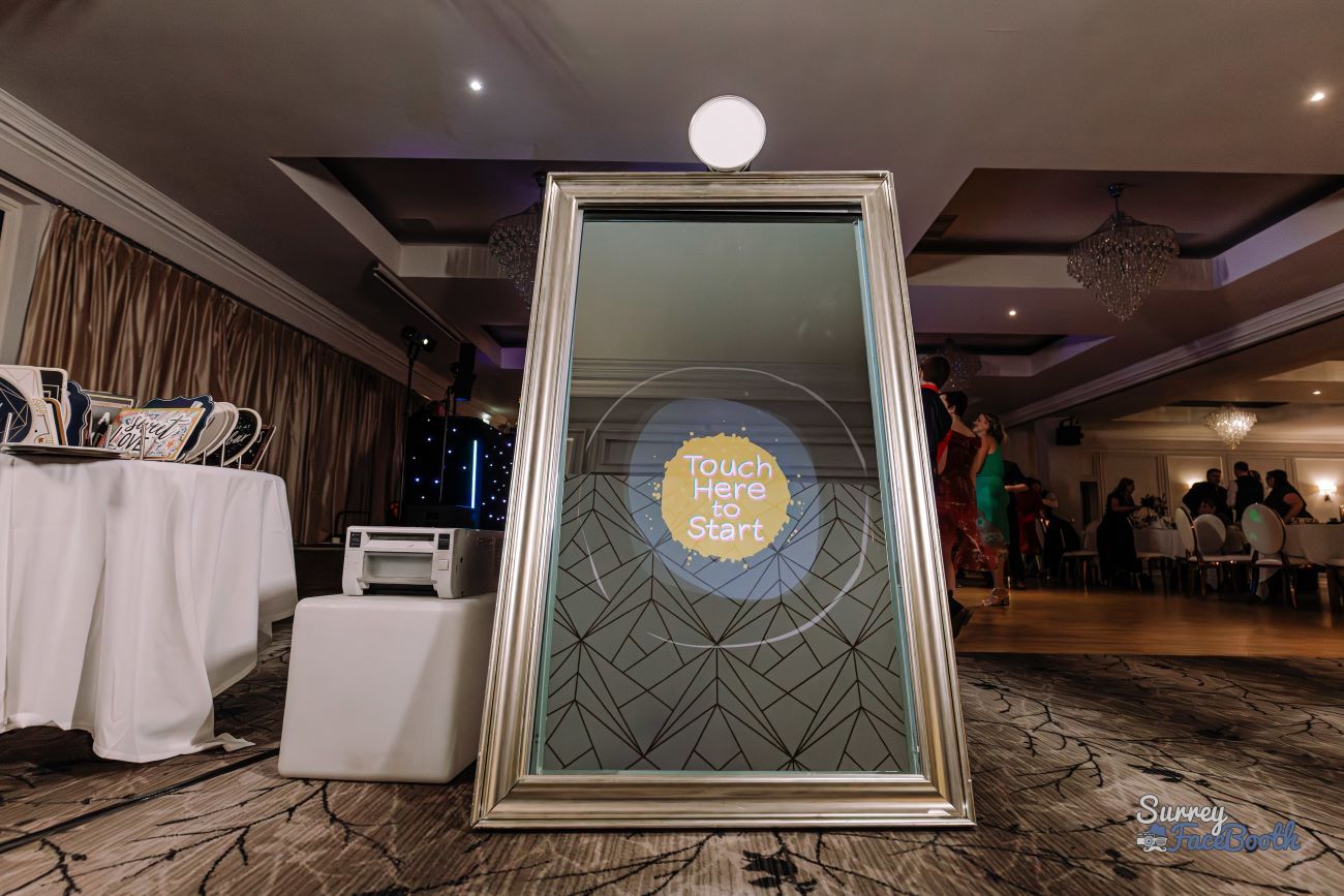 Silver-framed full-length magic mirror set up in a country-house hotel ballroom, 'Touch Here to Start' prompt on the screen, dye-sublimation printer on a side table and stacked photo albums beside it