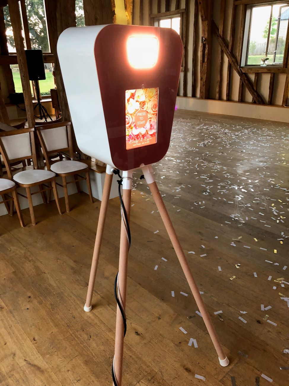 Retro pod set up in a rustic barn wedding venue with confetti scattered across the wooden floor, touchscreen showing a rose-floral 'Press to Start' overlay branded for the couple
