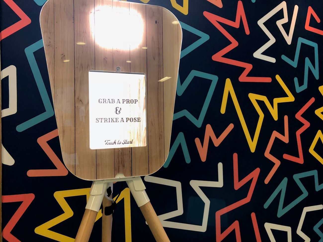 Front of the retro pod's wood-fascia panel — fill light at the top, 'Grab a Prop & Strike a Pose / Touch to Start' touchscreen graphic, against the Memphis-style geometric backdrop