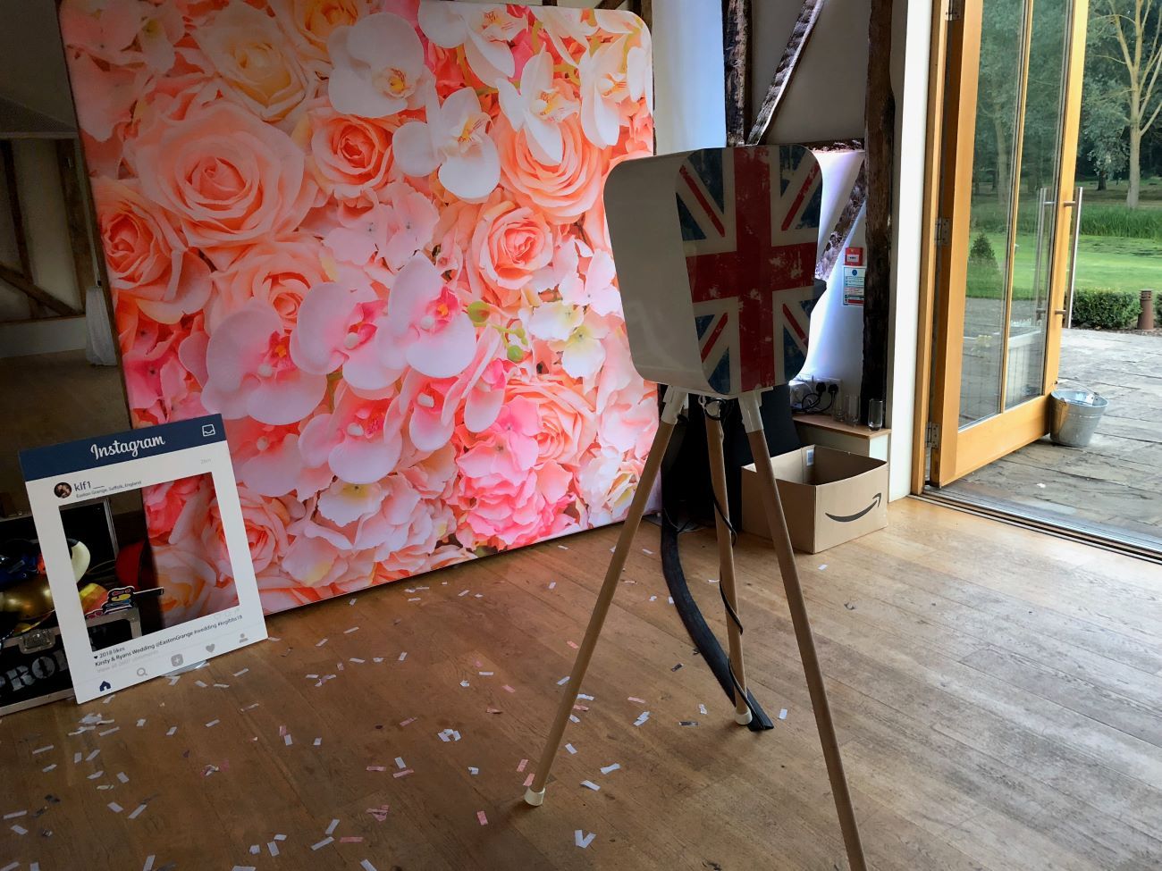 Retro pod on its wooden tripod next to a pink-and-white flower wall backdrop in a barn venue, with an Instagram-frame prop and confetti scattered on the floor