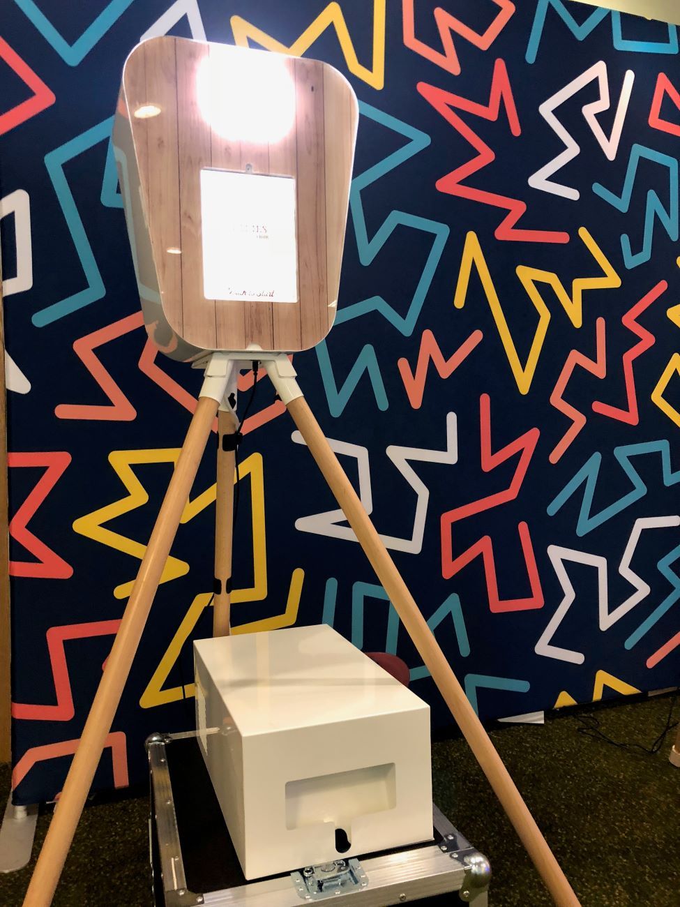 Retro pod photo booth on its wooden tripod against a Memphis-style geometric backdrop in navy with red, yellow, blue and white chevrons, fill light illuminated, separate white printer flight case on the floor in front