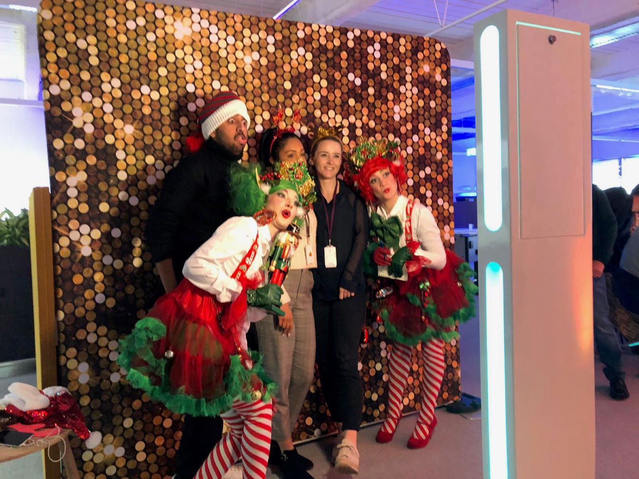 Corporate Christmas group photo at the selfie pod — guests in elf and nutcracker costumes posing in front of a gold sequin backdrop