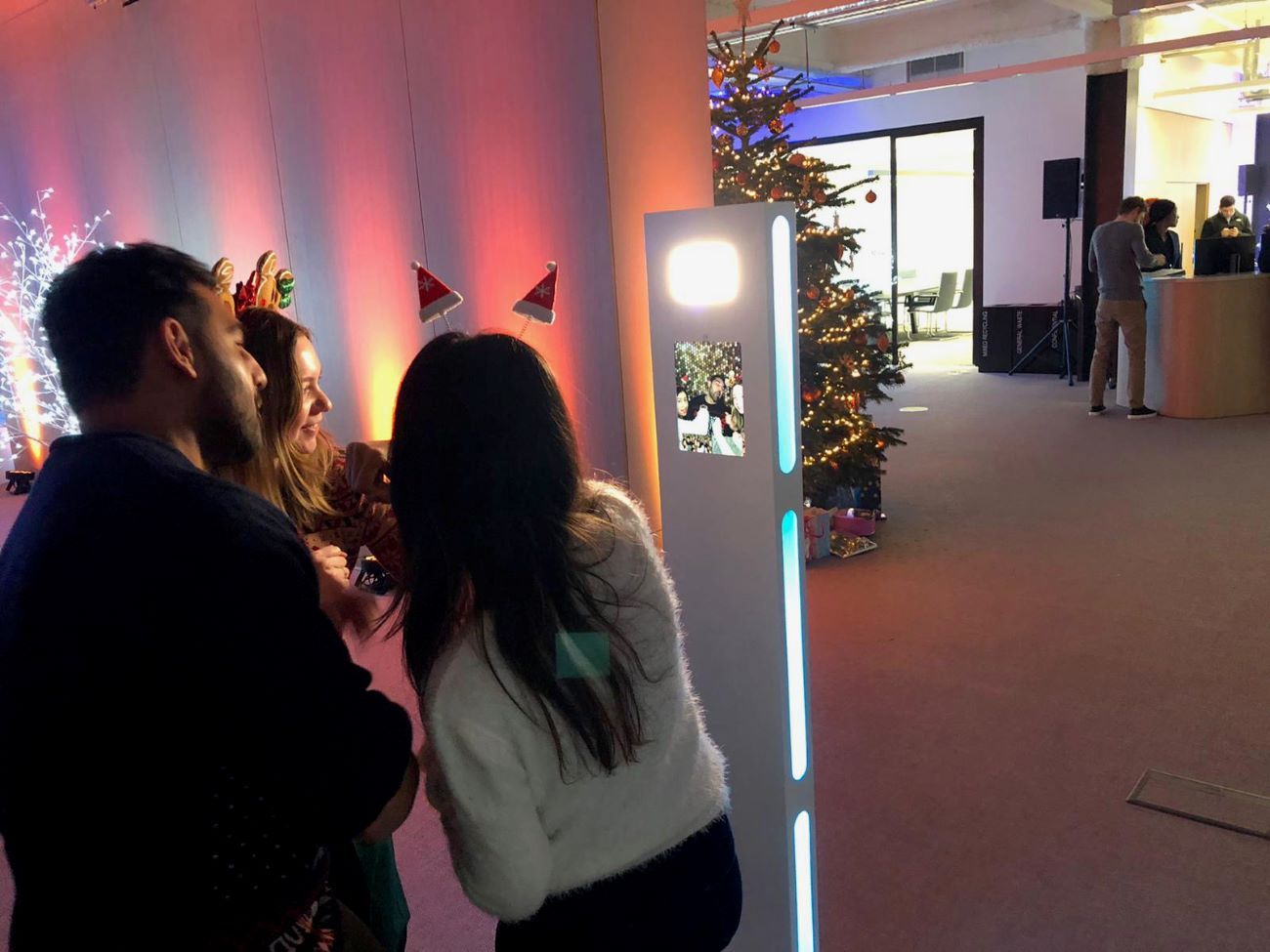 Three guests in Christmas reindeer and Santa hat headbands using the selfie pod at an office Christmas party, group preview showing on the touchscreen
