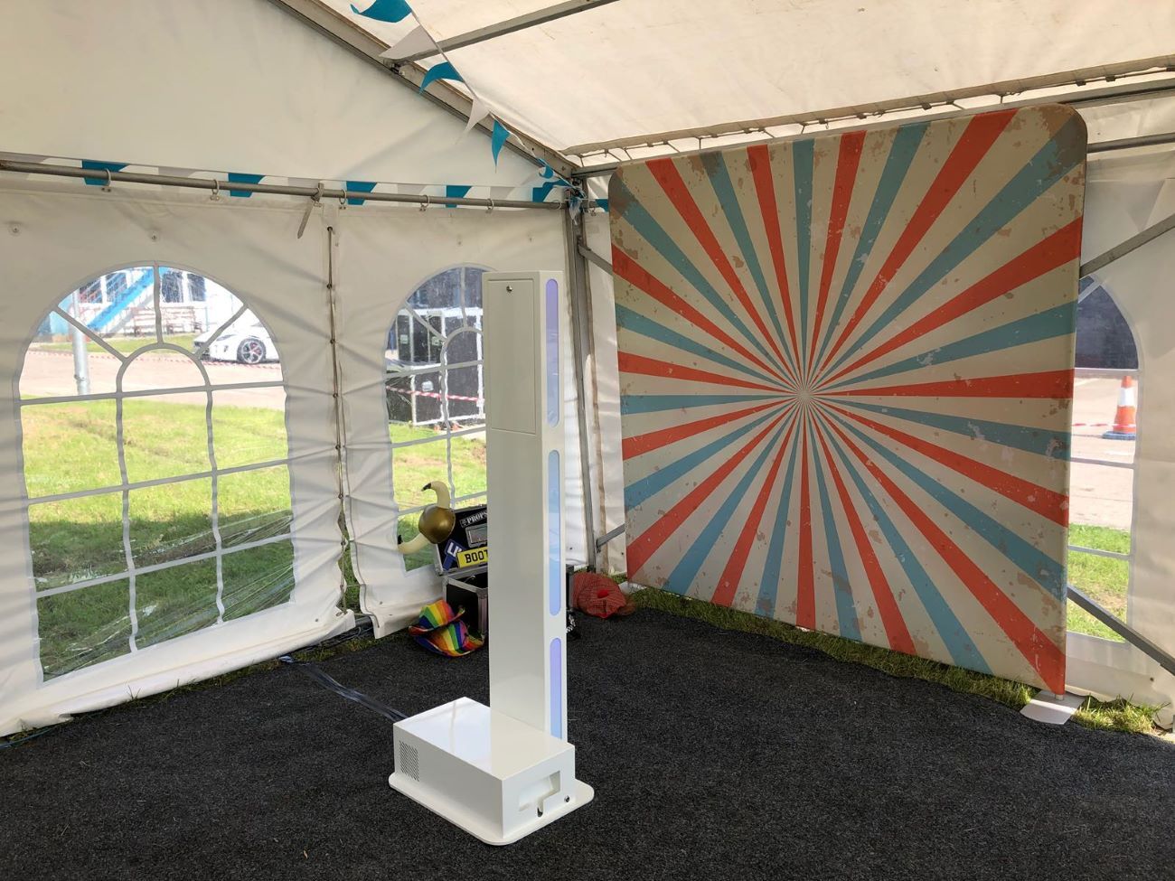 Selfie pod set up inside a garden marquee next to a red-and-cream sunburst backdrop, slim footprint visible against the marquee window