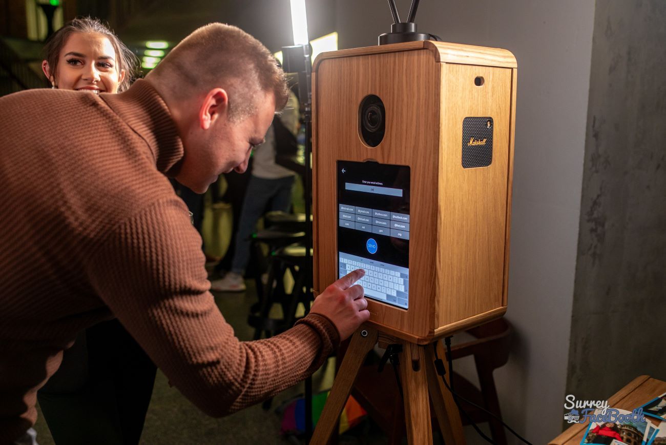 Guest at a corporate event tapping their email address into the vintage photo booth touchscreen to share their photo, with the booth's built-in Marshall speaker visible on the side