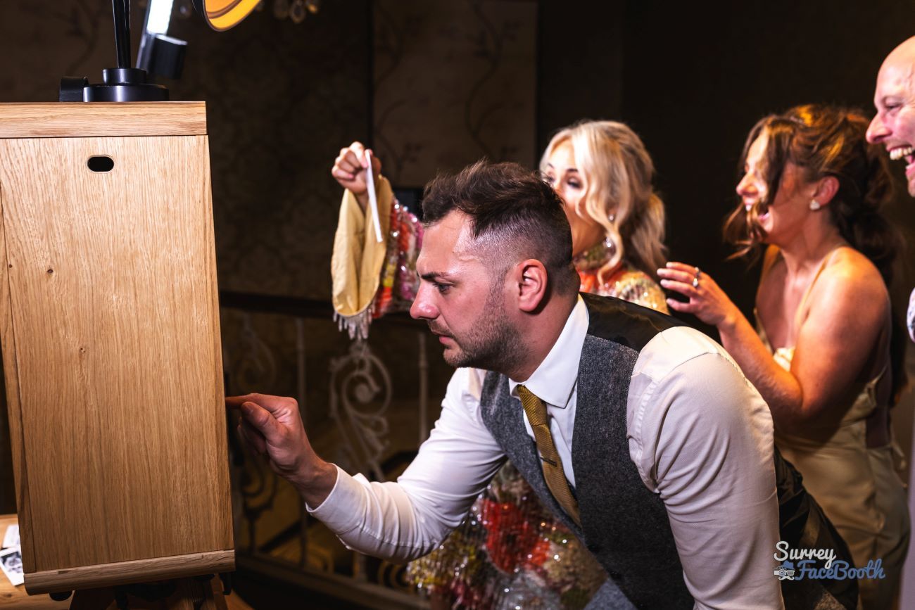 Wedding guest leaning in to tap the vintage photo booth touchscreen while the bride and other guests laugh in the background