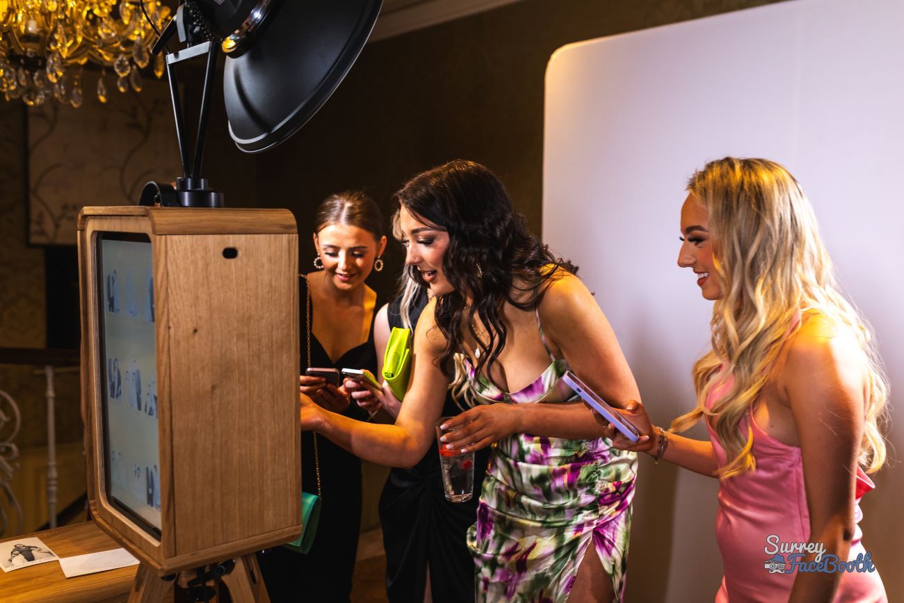 Three dressed-up wedding guests at a chandelier-lit venue gathered around the vintage photo booth, sharing their photos to their phones via QR code