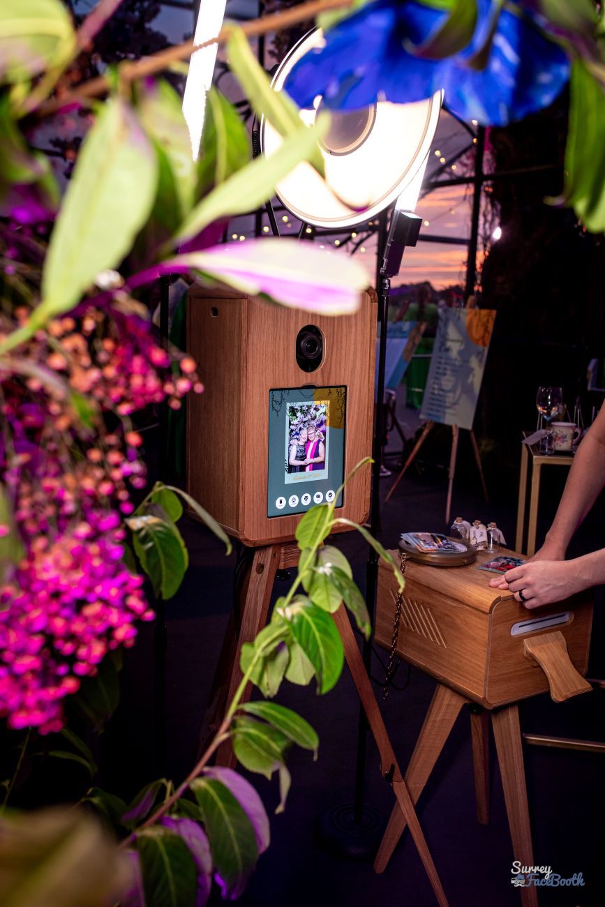 Vintage photo booth styled into a covered garden event setup — surrounded by hanging florals, fairy lights and a ring flash, screen showing a recent capture