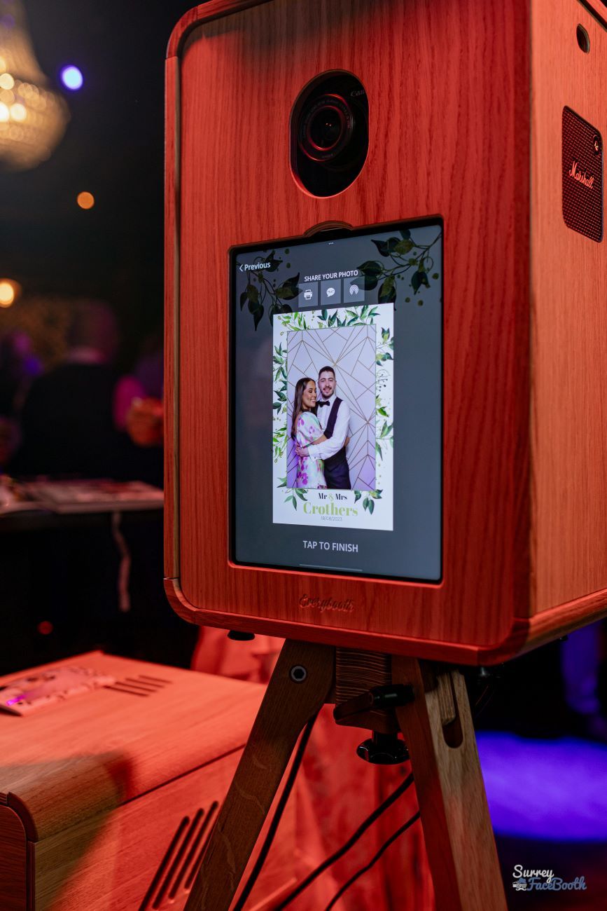 Red-finish variant of the vintage photo booth at a wedding, touchscreen showing the share-photo step with a green-foliage and monogram overlay for Mr and Mrs Crothers