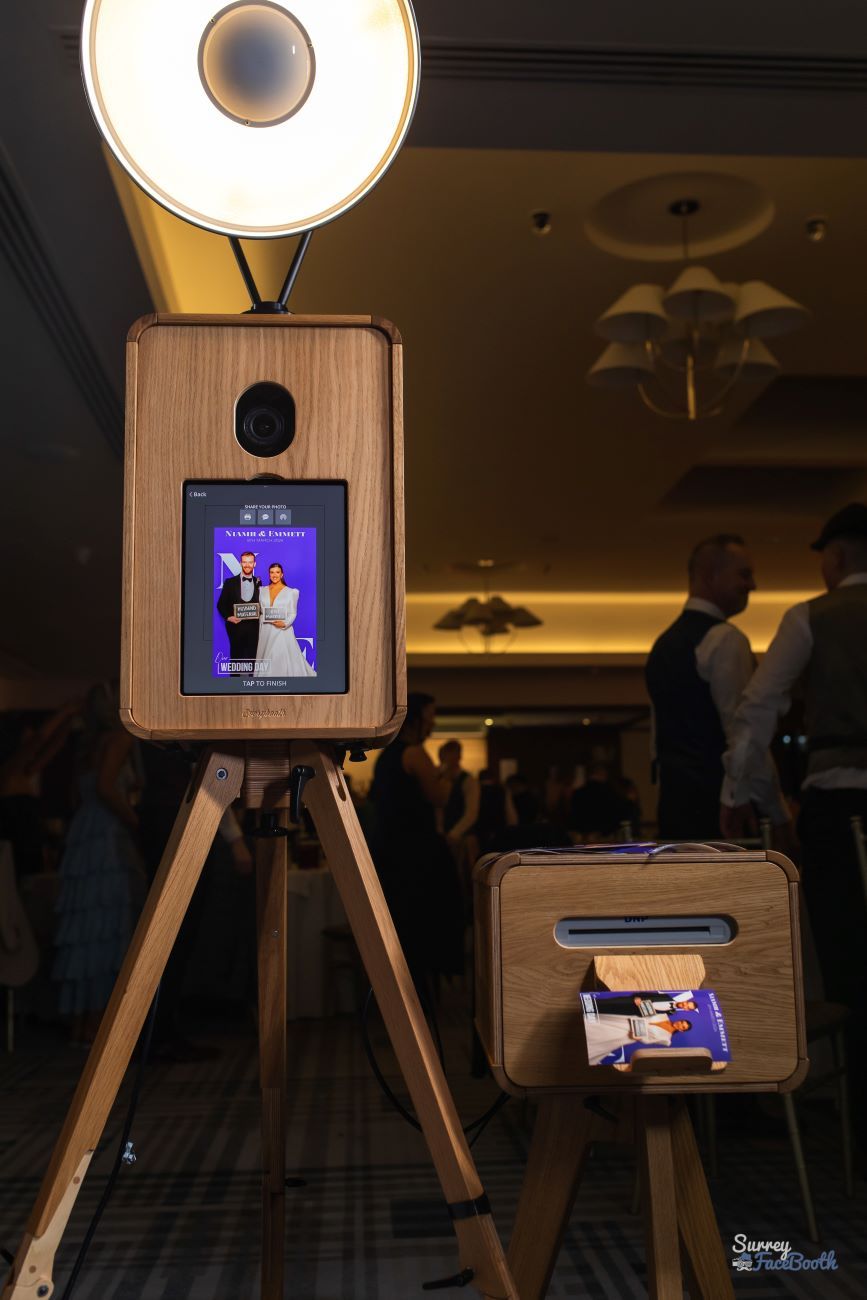Close-up of the vintage photo booth — large ring light overhead, touchscreen displaying a captured wedding-couple photo, the dye-sublimation printer below dispensing a fresh print