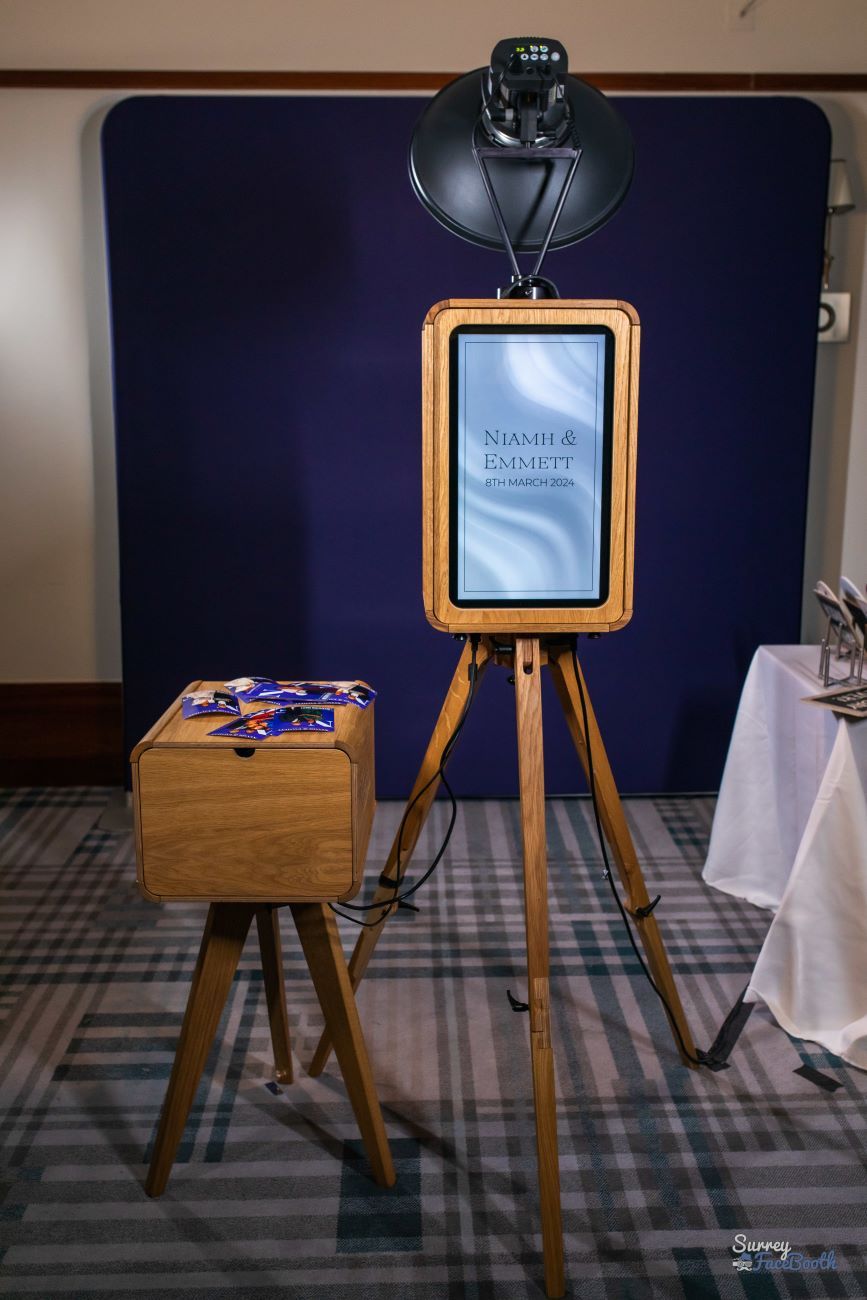 Vintage photo booth set up on a wooden tripod at a wedding venue, screen showing the couple's name and date with prints stacked on the matching wooden side cabinet