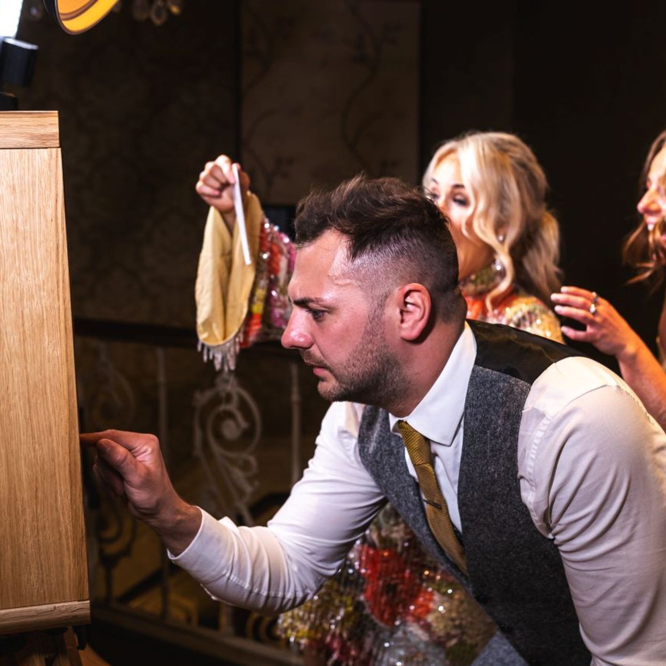 Wedding guest leaning in to tap the vintage photo booth touchscreen while the bride and other guests laugh in the background