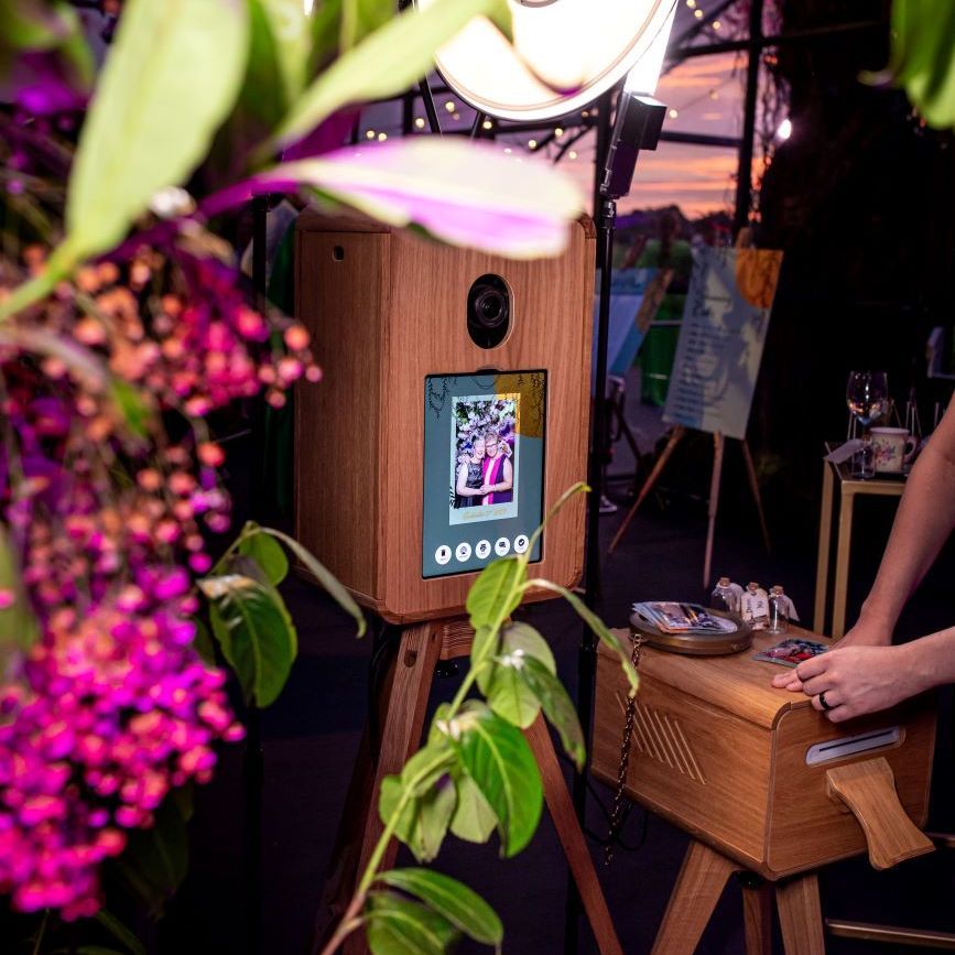 Vintage photo booth styled into a covered garden event setup — surrounded by hanging florals, fairy lights and a ring flash, screen showing a recent capture