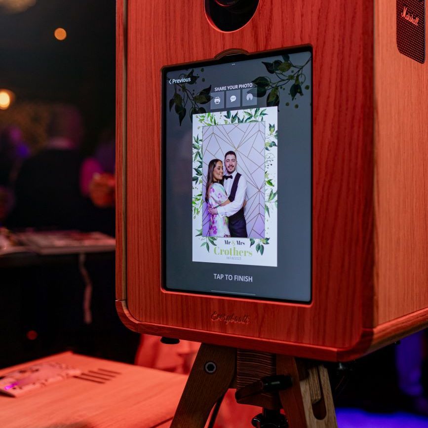 Red-finish variant of the vintage photo booth at a wedding, touchscreen showing the share-photo step with a green-foliage and monogram overlay for Mr and Mrs Crothers