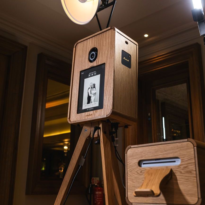 Vintage photo booth on its wooden tripod with the ring flash overhead, side cabinet housing the DNP dye-sublimation printer, set up in a chandelier-lit hotel ballroom for a wedding