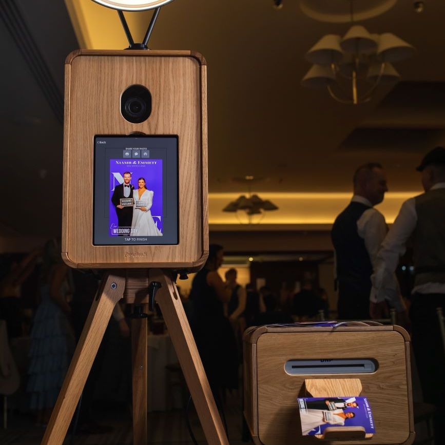 Close-up of the vintage photo booth — large ring light overhead, touchscreen displaying a captured wedding-couple photo, the dye-sublimation printer below dispensing a fresh print