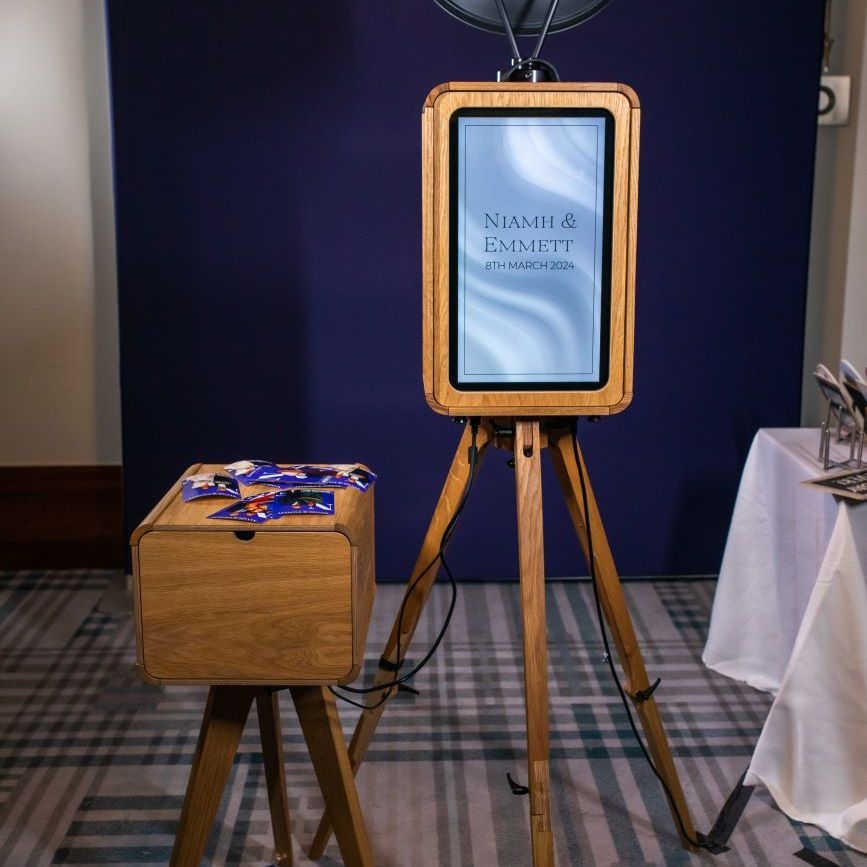 Vintage photo booth set up on a wooden tripod at a wedding venue, screen showing the couple's name and date with prints stacked on the matching wooden side cabinet